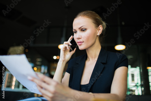 Serious business woman holding paper documents with contract and talking via mobile phone. Female successful employer reading resume and phoning via cell telephone. Leadership reading summary