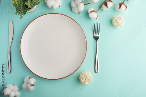 Empty white dish with knife and fork on a turquoise table background with a cotton branch, with copy space for your menu or recipe.Menu card for restaurants and table setting.Horizontal photo.Flat lay