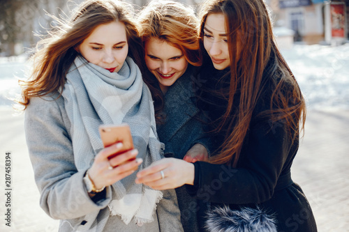 three beautiful elegant girls in winter coats walking together in a sunny winter city and use the phone