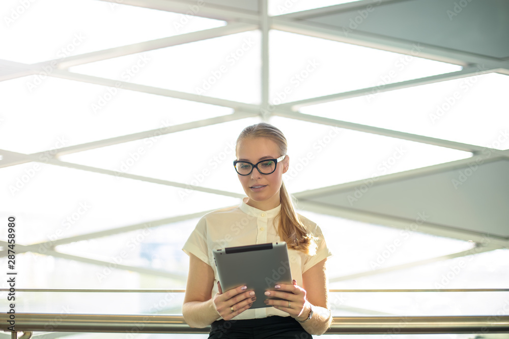 Proud woman entrepreneur in glasses and  formal wear reading e-mail on portable touch pad device while standing inside modern company interior. Female office worker online banking via digital tablet