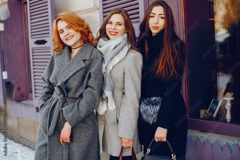 three beautiful elegant girls in winter coats standing together in a sunny winter city near building
