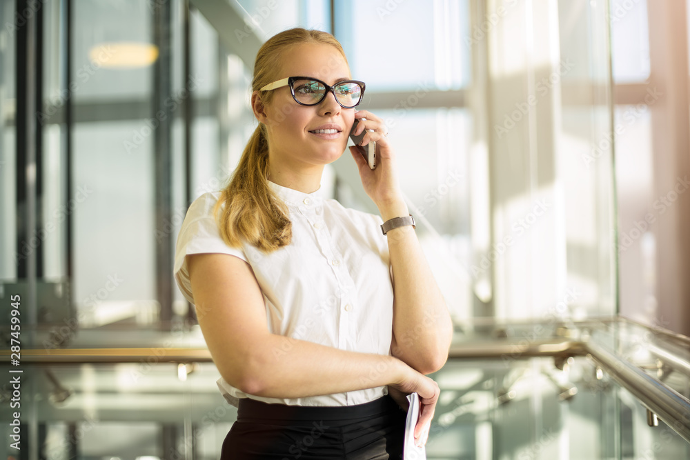Fototapeta premium Smiling woman in official wear successful government worker talking via smartphone while standing in modern interior during break at job. Joyful female in glasses skilled entrepreneur phoning