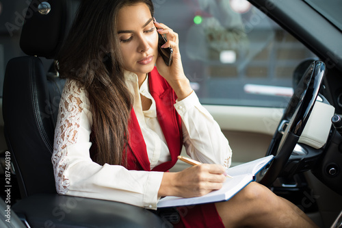 Gorgeous woman skilled office worker writing information in textbook and talking via mobile phone while sitting in luxury automobile. Confident female jurist having cell telephone conversation in car