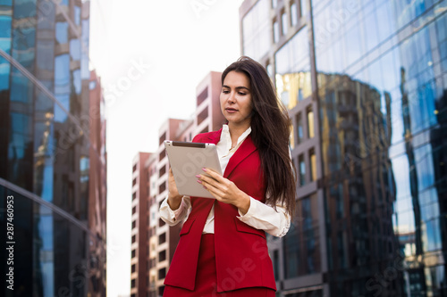 Successful woman professional real estate analyst using applications on modern touch pad while standing outdoors near company. Confident female broker checking e-mail on digital tablet