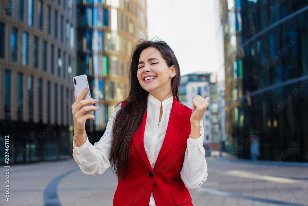 Cheerful smiling lucky woman managing director received on cell telephone happy good news in text message while standing outdoors near business centre during work break in enterprise