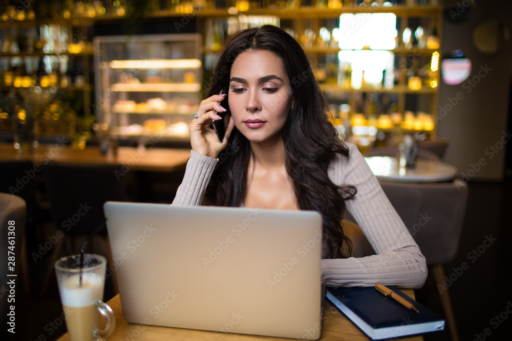 Confident business woman talking via mobile phone and reading e-mail on laptop computer while sitting in luxury restaurant interior Confident business woman talking via mobile phone and reading e-mail on laptop computer while sitting in luxury restaurant interior