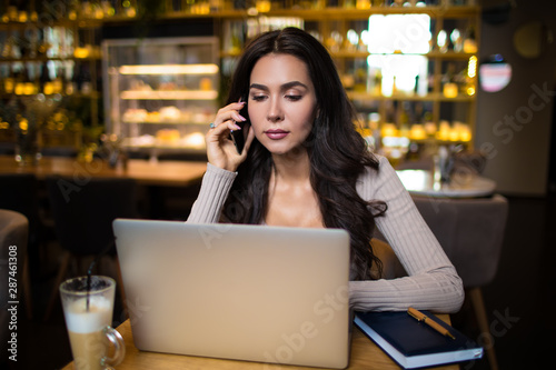 Confident business woman talking via mobile phone and reading e-mail on laptop computer while sitting in luxury restaurant interior 