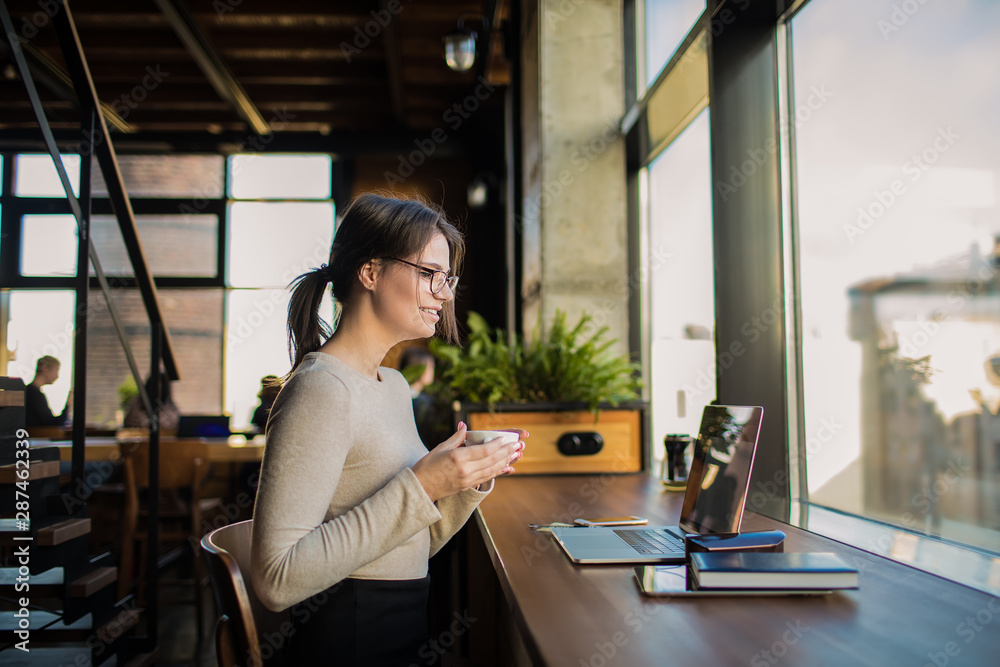 Smiling hipster girl in spectacles enjoying tea while watching movie via laptop computer, relaxing in coffee shop during recreation time . Cheerful female having online video conference via netbook