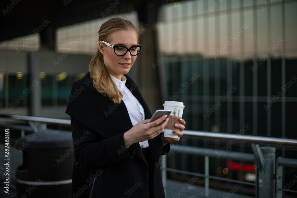 Woman in stylish spectacles checking notifications on mobile phone while standing with take away coffee outdoors against building with contemporary exterior. Female lawyer online payment via cellphone