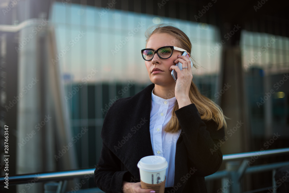 Serious woman confident business worker in fashionable spectacles and formal wear having smartphone conversation while standing with take away coffee outside enterprise building. Female leadership