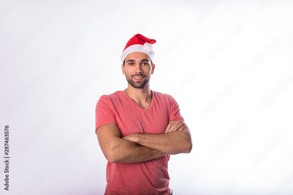 Portrait of a cheerful with good mood bearded handsome man in red Canta Clause hat looking in camera while standing with crossed arms isolated in studio against white background with copy space