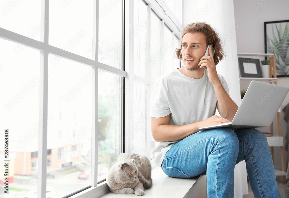 Man with laptop talking by phone while sitting on window sill with cute cat - obrazy, fototapety, plakaty 