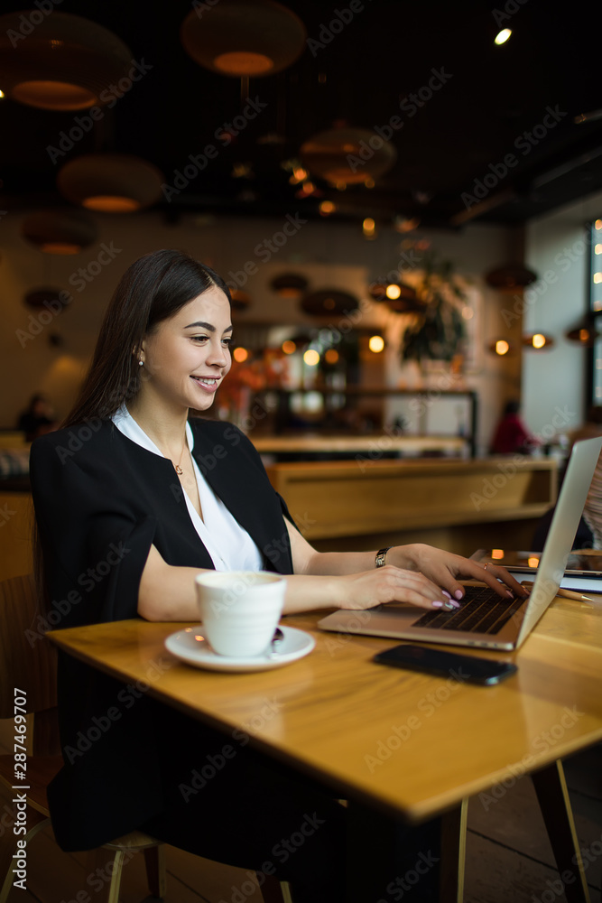 Smiling woman with good mood fashionable blogger typing promotional article on website via laptop computer while sitting in restaurant during recreation time in weekend. Online webinar via netbook Smiling woman with good mood fashionable blogger typing promotional article on website via laptop computer while sitting in restaurant during recreation time in weekend. Online webinar via netbook