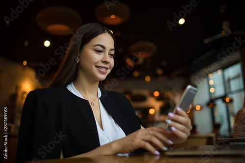 Attractive smiling woman reading pleasant text message on smartphone during leisure time in coffee shop. Happy joyful female manager watching video on cell telephone, sitting in restaurant