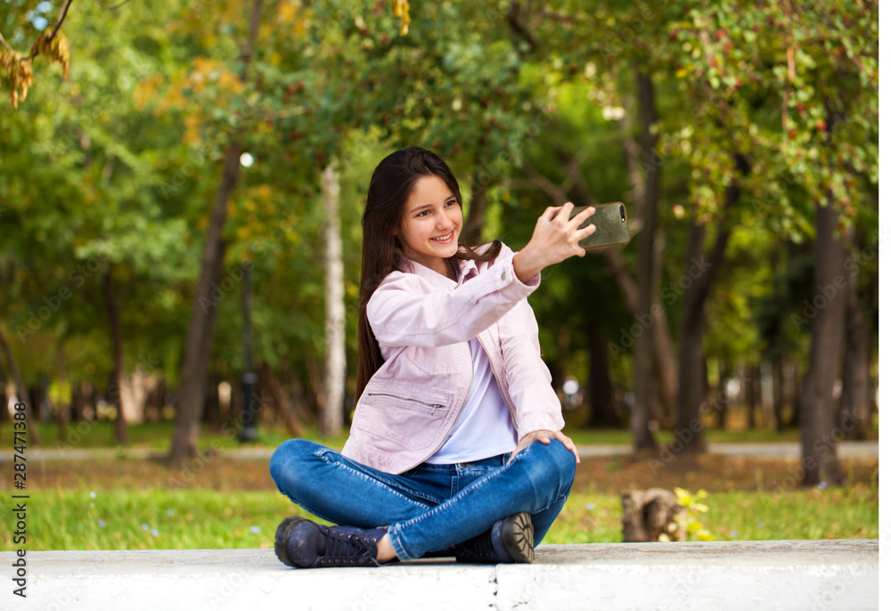Brunette girl photographs herself on a cell phone while sitting in an autumn park. Brunette girl photographs herself on a cell phone while sitting in an autumn park.