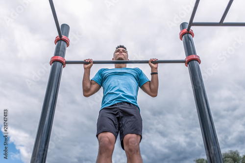 A male athlete pulls himself up on a horizontal bar in the summer in the city. Concept of strength, fitness workout in the city. Sportswear, blue t-shirt shorts, clouds background sports playground.
