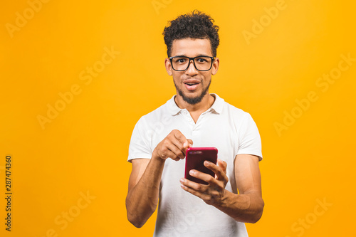 Smiling black young man in glasses holding phone isolated on yellow studio background with copy space aside, african guy using mobile applications, texting. © denis_vermenko