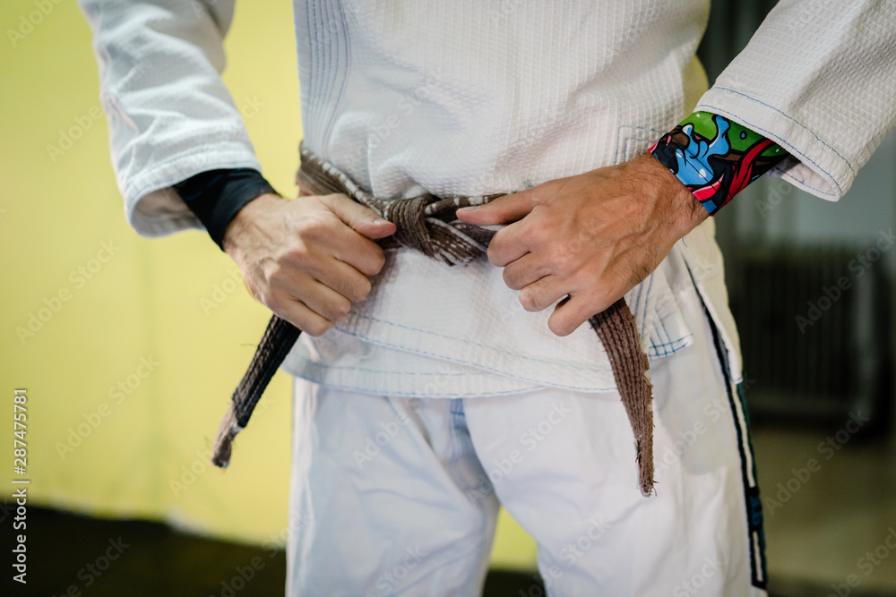 Close up on midsection of a man in martial arts gi kimono uniform tying his brown BJJ brazilian jiu jitsu belt around his waist in training at the academy