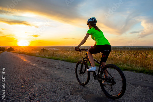 Beautiful girl cyclist riding a bike on the road towards the sunset.