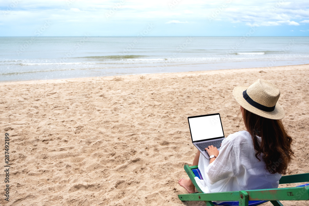 Mockup image of a woman using and typing on laptop computer with blank desktop screen while sitting on beach chair on the beach
