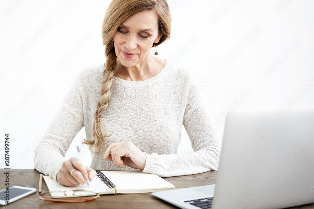 Horizontal shot of cheerful elegant sixty year old female teacher dressed in stylish sweater writing down in copybook while checking home assignments, smiling, using notebook pc and mobile phone Horizontal shot of cheerful elegant sixty year old female teacher dressed in stylish sweater writing down in copybook while checking home assignments, smiling, using notebook pc and mobile phone