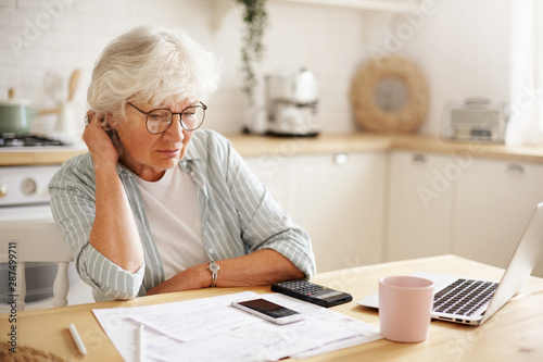 People, age, technology and finances. Depressed unhappy retired woman paying domestic bills online, trying hard to make both ends meet, sitting at kitchen table, surrounded with papers, using gadgets photo