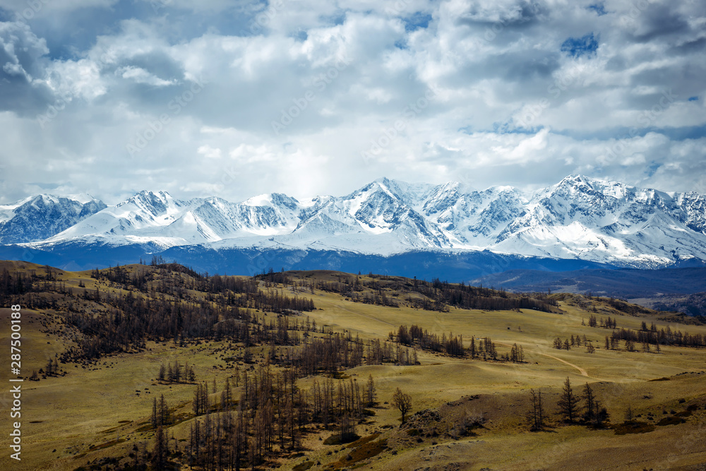 Fantastic mountain landscape. Rocky mountains with snowy peaks, hills covered with grass in the Alpine scene on a bright autumn day with blue sky and clouds. View of steppe and snow-covered mountains.