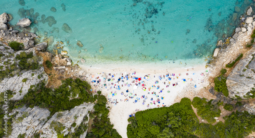 View from above, stunning aerial view of a beautiful beach full of beach umbrellas and people sunbathing and swimming on a turquoise water. Cala Gonone, Sardinia, Italy. View from above, stunning aerial view of a beautiful beach full of beach umbrellas and people sunbathing and swimming on a turquoise water. Cala Gonone, Sardinia, Italy.