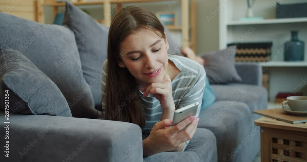 Pretty blond lady is using smartphone and smiling lying on couch in apartment looking at screen enjoying content. Entertainment, people and home concept. Pretty blond lady is using smartphone and smiling lying on couch in apartment looking at screen enjoying content. Entertainment, people and home concept.