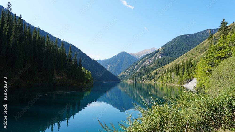 Kolsay lake located in the mountains of Kazakhstan. Turquoise water as a mirror. The water reflects the mountains, green hills, tall spruce, grass, sky and clouds. Beautiful landscape of mountain lake