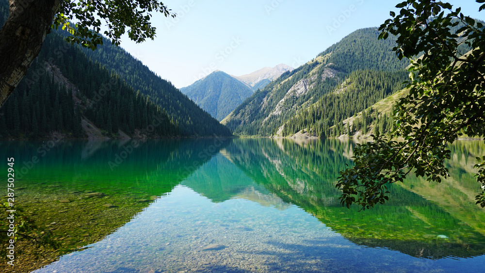 Kolsay lake located in the mountains of Kazakhstan. Turquoise water as a mirror. The water reflects the mountains, green hills, tall spruce, grass, sky and clouds. Beautiful landscape of mountain lake