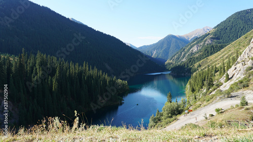 Turquoise water mirror mountain lake Kolsai. The boat slowly floats on the mirror surface, everything is reflected. It offers views of green mountains, spruce, trees, grass and hills. Romantic setting