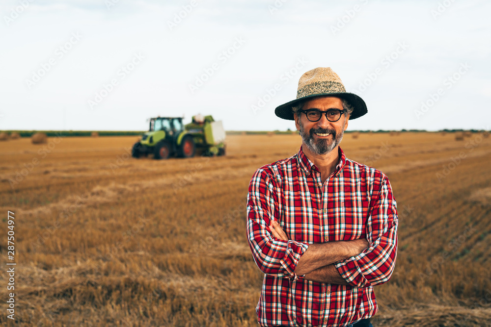 portrait of middle aged rancher standing crossed arms outdoors, agricultural machine in blurred background