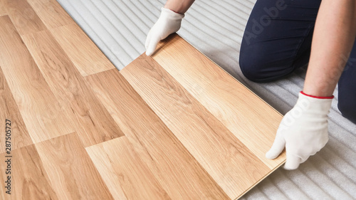 Installing laminated floor, detail on worker hands in white gloves fitting wooden tile, over white foam base layer