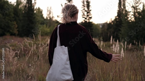 Girl walks in the garden or park, touches spikelets at summer evening . Trendy hipster girl with dreads enjoying nature. View from the back photo