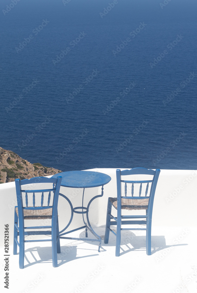Greece, the island of Sikinos. Two traditional taverna chairs and a blue table. In the background, the  blue Aegean sea. Picture taken at a cafe high above the waters. Peace, simplicity and nature.