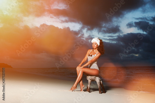 Fashion woman sit on the sunny beach sunset background. Happy lifestyle. White sand, dawn cloudy sky and blue sea of tropical beach. Sexy tanned woman on the summer beach.