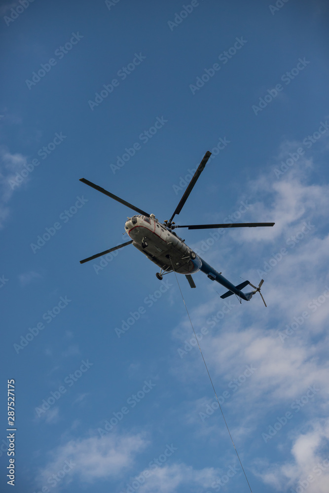A white and blue helicopter flying in the blue sky - the cable hangs out of the helicopter A white and blue helicopter flying in the blue sky - the cable hangs out of the helicopter