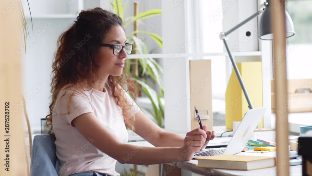 Tracking right of Caucasian businesswoman with long curly hair and eyeglasses sitting at desktop in office, working then looking at camera and smiling Tracking right of Caucasian businesswoman with long curly hair and eyeglasses sitting at desktop in office, working then looking at camera and smiling