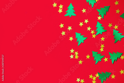 Green metallic foil christmas trees and gold stars confetti sparse on red background. Simple holiday concept. Design template. Frame with copy space. Winter festive backdrop. Top view, flat lay.