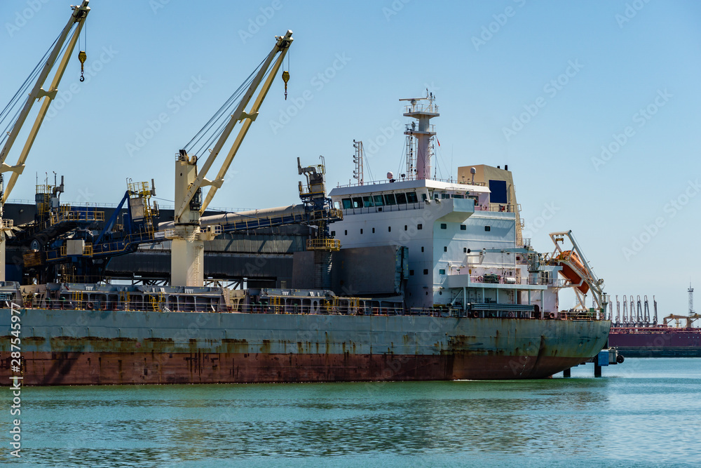 Cargo ship is equipped with cranes for loading. Close-up. Cargo ship stands at pier in port of Tuapse.  Sea is calm. Sea water of emerald color. Cargo ship occupies entire length of large berth.