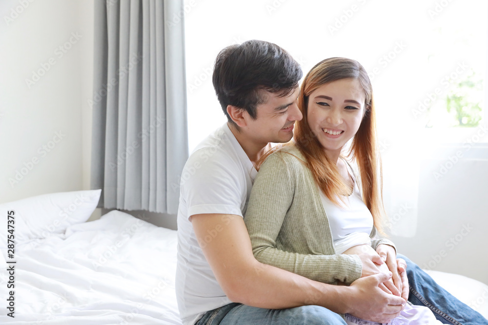 close up young caucasian husband embracing young pregnant asian belly wife on bed in bedroom
