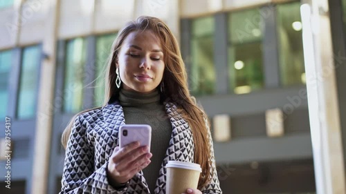 Smiling young European woman in gray coat using smartphone and holding disposable coffee cup in city street. Concept of communication. Slider slow motion medium shot
