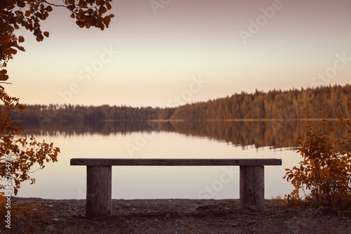 On the shore of the lake is a bench. Autumn forest background is blurred