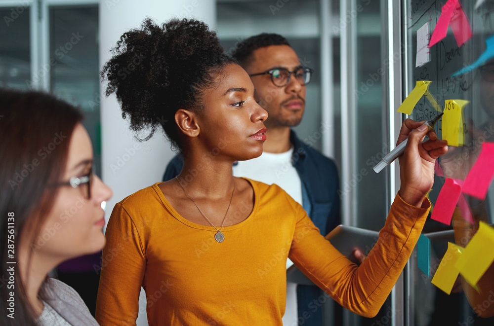 © StratfordProductions - Young creative businesswoman standing with her colleagues writing new ideas on sticky notes over glass wall