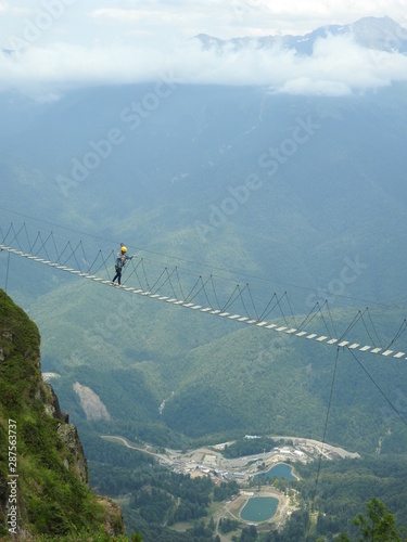 a tourist walks over a precipice along a thin and narrow suspension cable bridge against the backdrop of a mountainous Caucasian landscape. View on a summer sunny day