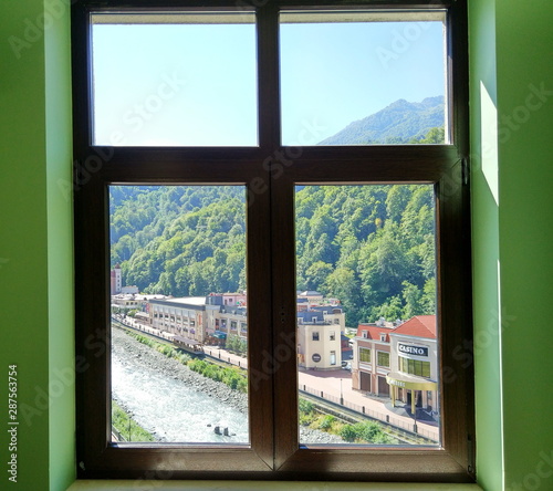Hotel window on ski resort in summer season - view of neighboring hotels, mountain river and mountains