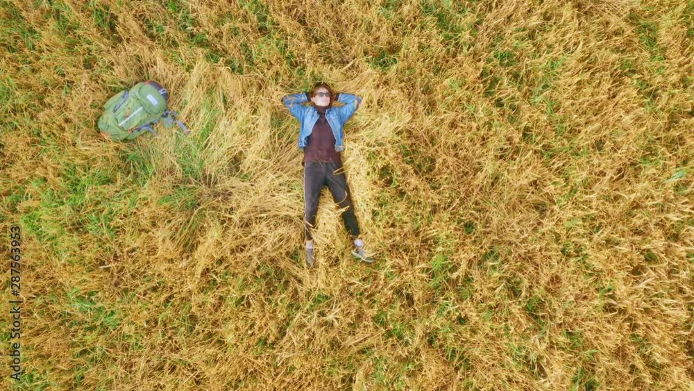 A young girl, a traveler and a hitchhiker, lies next to a backpack on a cereal field sown with wheat or rye. Summer sunny day. Aerial top view. A young girl, a traveler and a hitchhiker, lies next to a backpack on a cereal field sown with wheat or rye. Summer sunny day. Aerial top view.