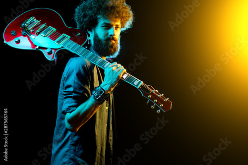 Portrait of hipster man with curly hair with red guitar in neon lights.