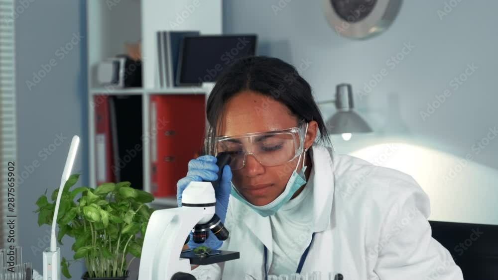 Multiracial research scientist getting shocked with the result of the experiment seen under the microscope. Woman is in safety glasses and lab coat. Multiracial research scientist getting shocked with the result of the experiment seen under the microscope. Woman is in safety glasses and lab coat.